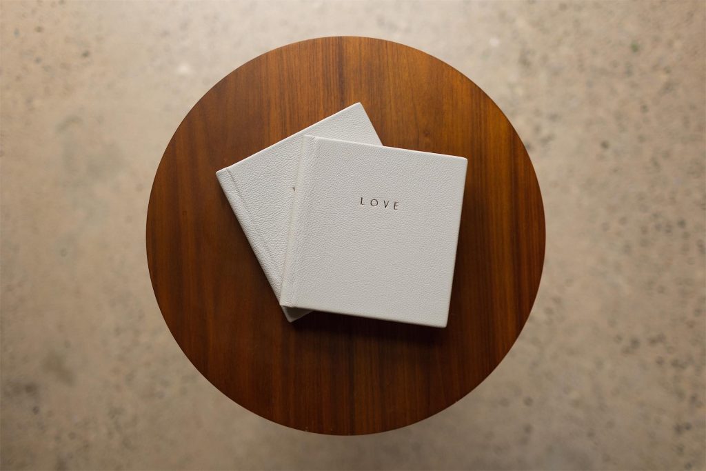 Two smaller copies of a wedding album stacked on a table with the word LOVE gold foiled on the grey leather cover