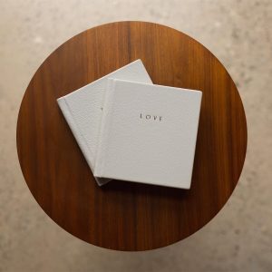 Two smaller copies of a wedding album stacked on a table with the word LOVE gold foiled on the grey leather cover