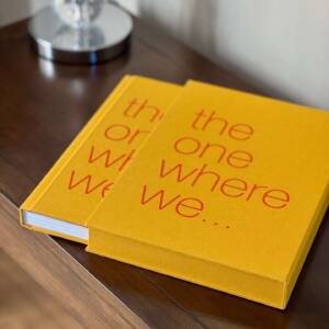 A coffee table book partly removed from its slipcase with an orange-coloured cover and red text reading 'the one where we ...'