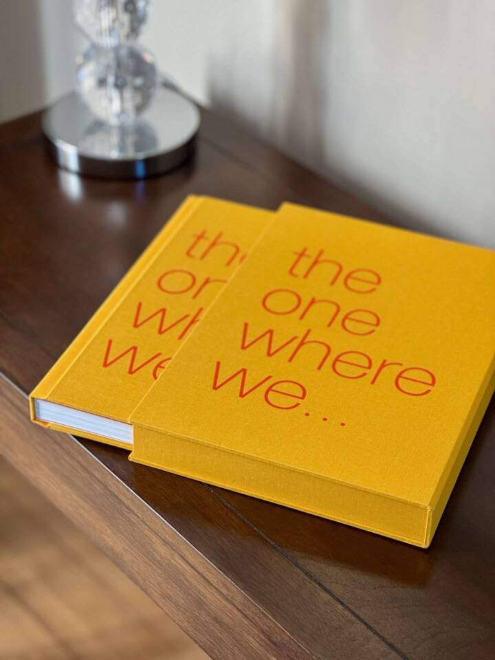 A coffee table book partly removed from its slipcase with an orange-coloured cover and red text reading 'the one where we ...'