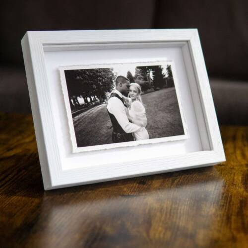 A frame on a wooden table with a fine art print of a groom cuddling the bride. The print is float-mounted in a frame with a shadow cast by the deckle edge of the 6x4" print in the 8x6" frame